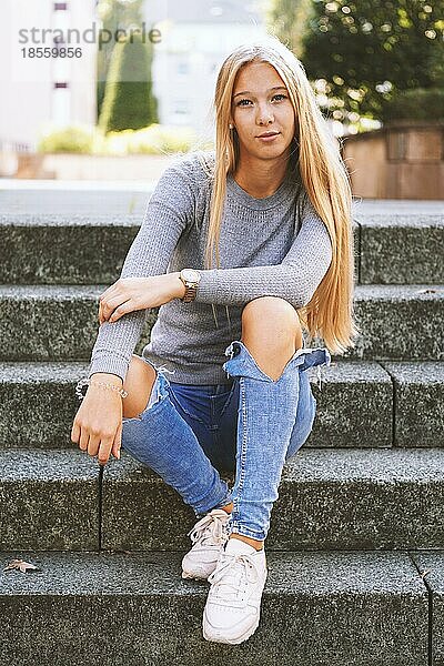 Teenager-Mädchen mit langen blonden Haaren und ausgebeulten Jeans sitzt draußen auf den Stufen - urbaner Lebensstil oder Straßenmode-Konzept