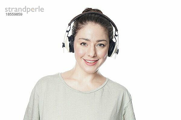 Glückliche junge Frau in den 20ern  die mit schnurlosen Over-Ear-Kopfhörern Musik hört - Mädchen vor weißem Hintergrund