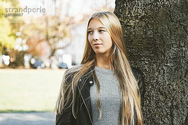Verträumtes Teenager-Mädchen mit langen blonden Haaren und Lederjacke lehnt an einem sonnigen Frühlingstag an einem Baum