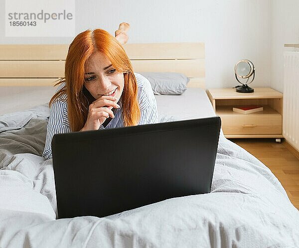 Glückliche junge Frau liegt mit Laptop auf dem Bett und entspannt sich zu Hause