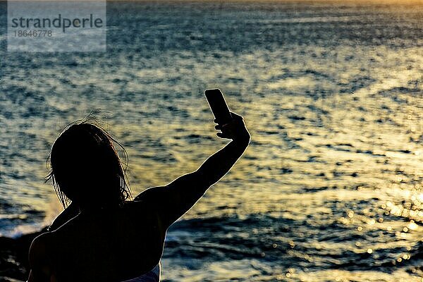 Silhouette eines Mädchens mit Smartphone  das im Sommer während des Sonnenuntergangs in der Stadt Salvador  Bahia  Brasilien  ein Selfie macht  Südamerika