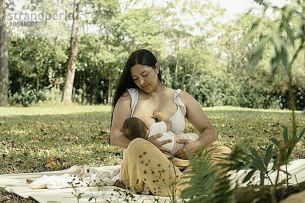 Die Mutter stillt ihr Baby  und beide wirken entspannt und zufrieden. Im Hintergrund sind Bäume und Grünflächen zu sehen  die eine friedliche und natürliche Atmosphäre schaffen