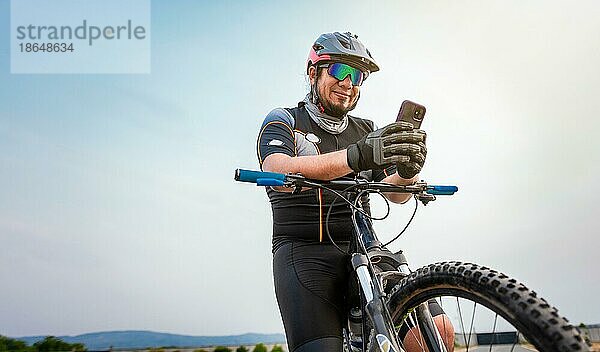 Männlicher Radfahrer mit Helm und Mobiltelefon. Radfahrer auf seinem Fahrrad SMS mit Telefon im Freien. Radfahrer Männchen mit Helm und Brille SMS auf Handy im Freien