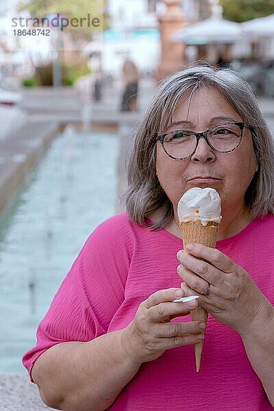 Nahaufnahme einer weißhaarigen Frau mit Brille  die vor einem Springbrunnen Eis isst