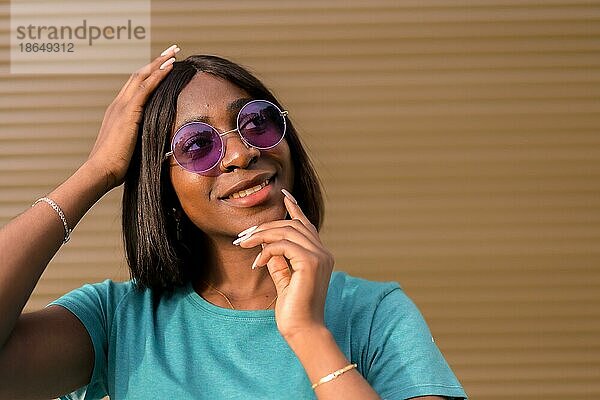 Sonnige Tage und gute Laune: Das Porträt einer jungen schwarzen Touristin mit grünem TShirt und Sonnenbrille vor einer braunen Wand