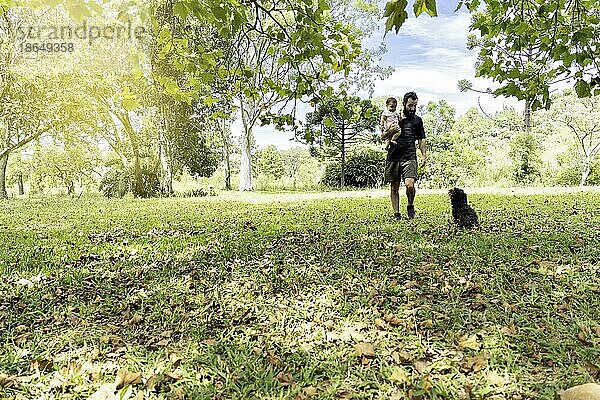 Vater hält sein Baby zu Fuß mit seinem Hund in das Feld von Bäumen umgeben. Vatertag Konzept