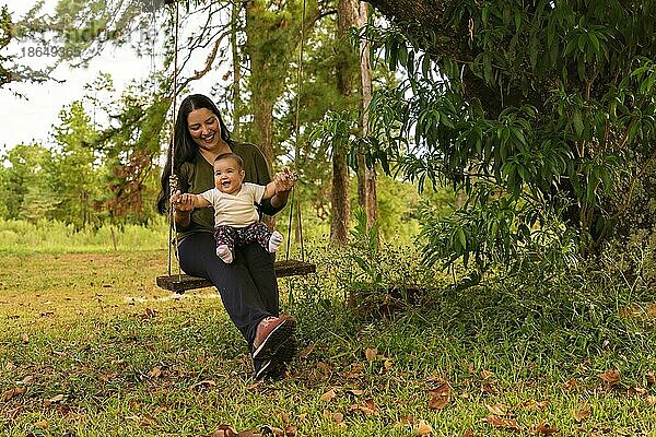 Glücklich fröhlich niedlichen Baby Mädchen sitzt auf einer Schaukel mit ihrer Mutter spielen auf dem Spielplatz