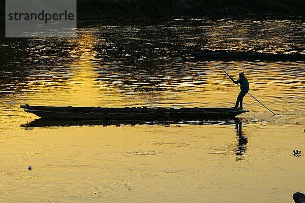 Bootsführer  der den Rapti Fluss in einem traditionellen Einbaum aus Holz überquert. Das Boot wird bewegt  indem man eine lange Stange gegen den Boden des Flusses stößt  wie hier zu sehen ist. In der Region werden die Boote gewöhnlich aus einfachen Baumstämmen aus Seidenbaumwolle hergestellt und sind weit verbreitet. Der Rapti Fluss (East Rapti River) bildet die nördliche Grenze des Chitwan Nationalparks  der zum UNESCO Welterbe gehört. Ein Blick auf den Sonnenuntergang  mit der Silhouette des Mannes im Gegenlicht der untergehenden Sonne  die sich im Wasser spiegelt. Dorf Sauraha  Bezirk Chitwan