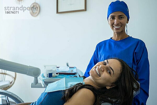Schöne junge Frau lächelt auf dem Zahnarztstuhl. Sie und ihr Arzt schauen zufrieden mit dem Ergebnis der kieferorthopädischen Behandlung in die Kamera