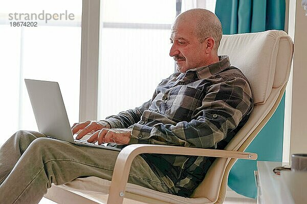 Mann mit Schnurrbart  kariertem Hemd und Jeans sitzt in einem Sessel in seinem Wohnzimmer und benutzt seinen Laptop