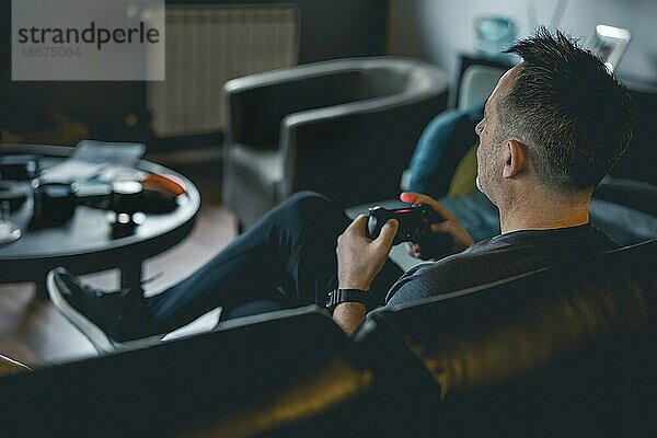 Ein erwachsener Mann sitzt auf einem Sofa und hält einen Joystick  während er ein Videospiel auf einer Konsole spielt