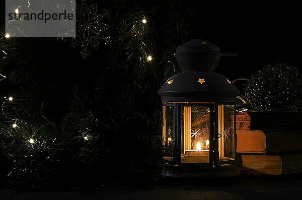 Weiße Laterne mit einer brennenden Kerze. Bücher  Weihnachtsbaum Zweig und Lichter auf dem Hintergrund. Nacht dunkles Bild. Weihnachten Geheimnis Stimmung