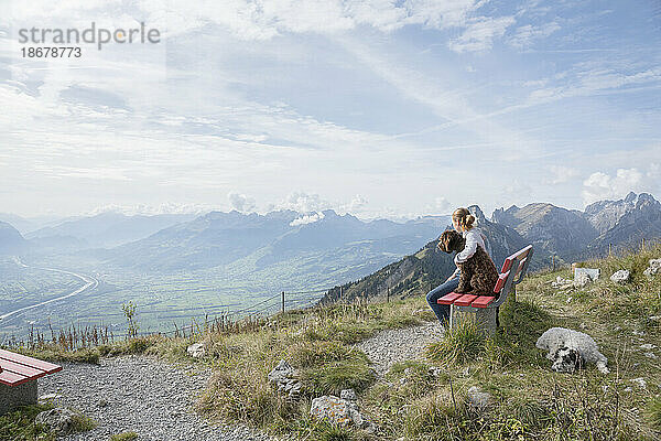 Mädchen mit Hunden sitzt auf einer Bank und genießt die malerische  majestätische  sonnige Aussicht auf die Bergkette der Schweiz