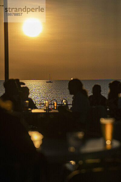 Silhouetten von Menschen  die bei Sonnenuntergang Bier auf der Terrasse am Wasser trinken und auf das Meer blicken