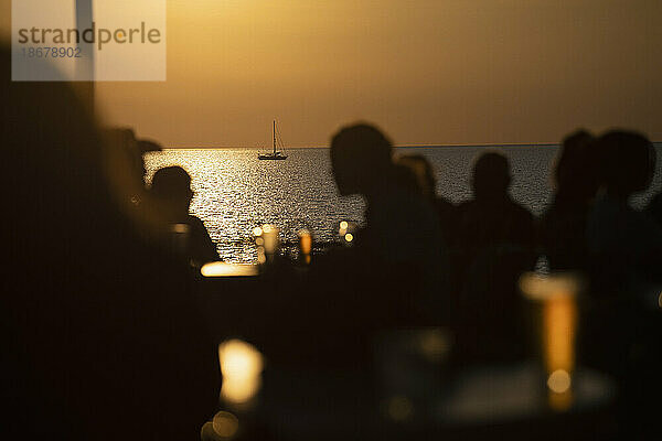 Silhouetten von Menschen  die bei Sonnenuntergang Bier auf der Terrasse am Meer trinken