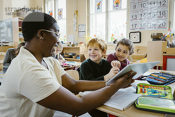 Glücklicher Lehrer mit digitalem Tablet unterrichtet Junge und Mädchen sitzen am Schreibtisch im Klassenzimmer