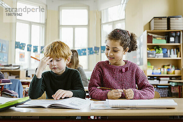 Lächelndes Mädchen sitzt neben einem verwirrten blonden Jungen am Schreibtisch im Klassenzimmer