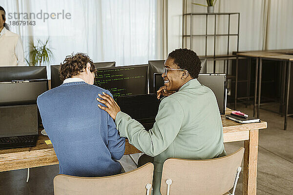 Rückansicht einer lächelnden Programmiererin  die mit einem männlichen Kollegen an einem Computertisch im Büro sitzt