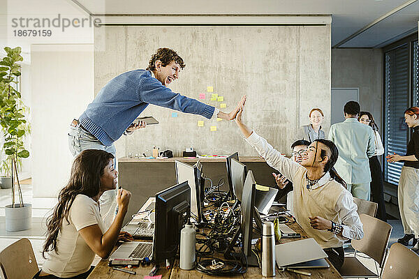Fröhliche Unternehmer geben sich die Hand  während Kollegen in einem kreativen Büro am Schreibtisch sitzen