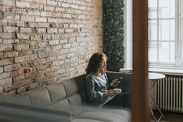 Geschäftsfrau  die während der Kaffeepause im Büro auf dem Sofa sitzend ihr Smartphone benutzt