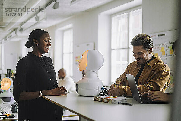 Multirassische lächelnde Studenten mit beleuchtetem sozialen Roboter am Schreibtisch im Innovationslabor