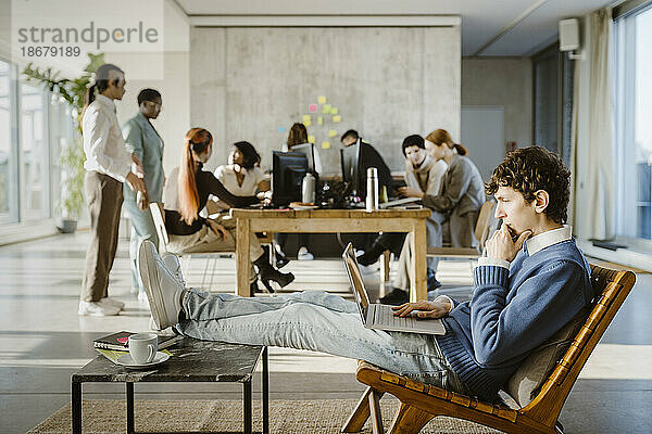 Seitenansicht eines jungen Geschäftsmannes  der auf einem Stuhl einen Laptop benutzt  während im Hintergrund Kollegen im Kreativbüro zu sehen sind