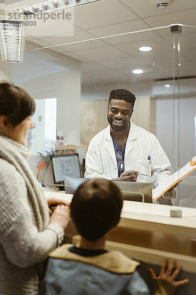 Lächelnder männlicher Rezeptionist im Gespräch mit Mutter und Sohn durch einen transparenten Schild in einer Klinik