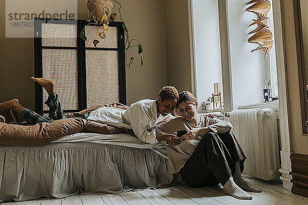 Lächelndes nicht-binäres Paar  das sich ein Smartphone teilt  während es seine Freizeit im Schlafzimmer zu Hause verbringt