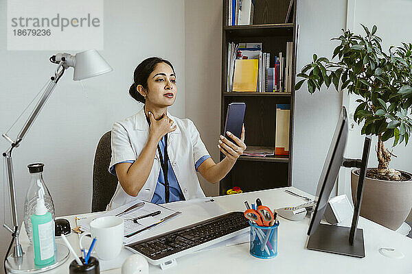 Ärztin bei der Aufklärung eines Patienten per Online-Videoanruf über das Mobiltelefon  während sie am Schreibtisch in der Klinik sitzt