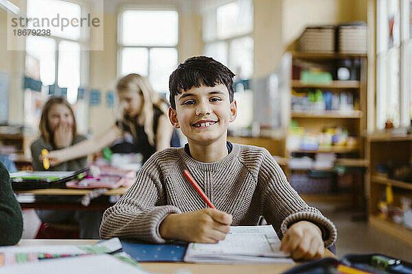 Porträt eines glücklichen männlichen Schülers  der am Schreibtisch im Klassenzimmer sitzt