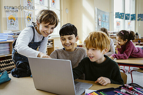 Glückliche Schüler  die gemeinsam einen Laptop am Schreibtisch im Klassenzimmer benutzen