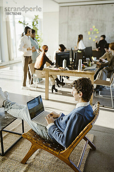 Junger kreativer Geschäftsmann  der einen Laptop benutzt  während er auf einem Stuhl im Kreativbüro sitzt