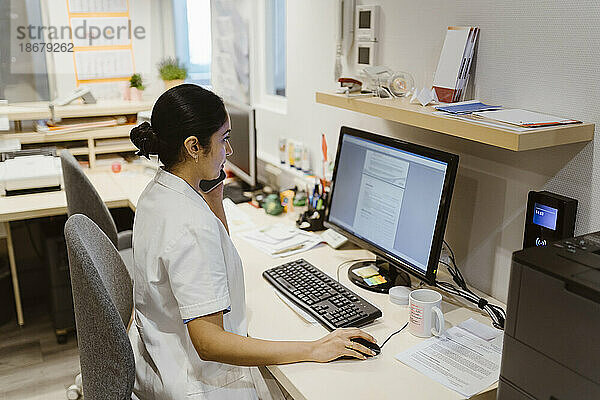 Mitarbeiterin des Gesundheitswesens benutzt einen Computer  während sie in einer Klinik mit einem Smartphone telefoniert
