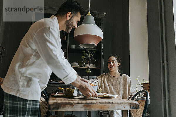 Lächelnder Mann  der das Frühstück mit einer nicht-binären Person serviert  die am Esstisch in einem Haus sitzt
