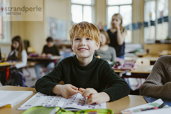 Lächelnder männlicher Schüler mit blondem Haar sitzt am Schreibtisch im Klassenzimmer