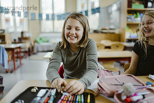Glückliches Schulmädchen  das lachend mit einem Freund am Schreibtisch im Klassenzimmer sitzt