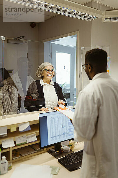 Lächelnde ältere Frau im Gespräch mit einem männlichen Rezeptionisten durch einen transparenten Schild in einer Klinik