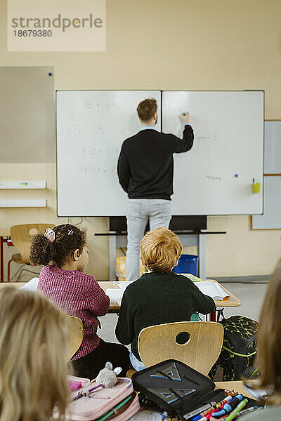 Rückansicht eines männlichen und eines weiblichen Schülers mit einem unterrichtenden Lehrer im Hintergrund