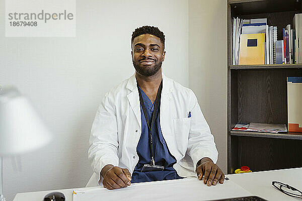 Porträt eines lächelnden männlichen Mitarbeiters im Gesundheitswesen  der einen Laborkittel trägt und am Schreibtisch in einer Klinik sitzt
