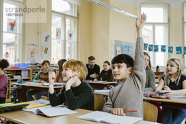 Junge mit erhobener Hand  der während einer Vorlesung antwortet  während er neben einem männlichen Freund im Klassenzimmer sitzt