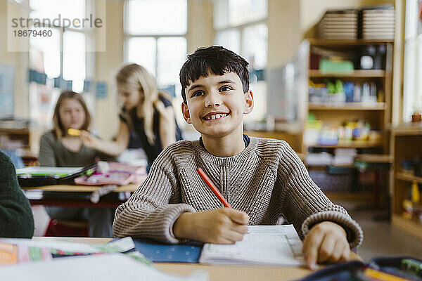 Lächelnder männlicher Schüler  der einen Bleistift hält  während er am Schreibtisch im Klassenzimmer sitzt
