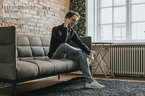 Lächelnder Geschäftsmann  der mit seinem Smartphone spricht  während er auf dem Sofa im Büro sitzt