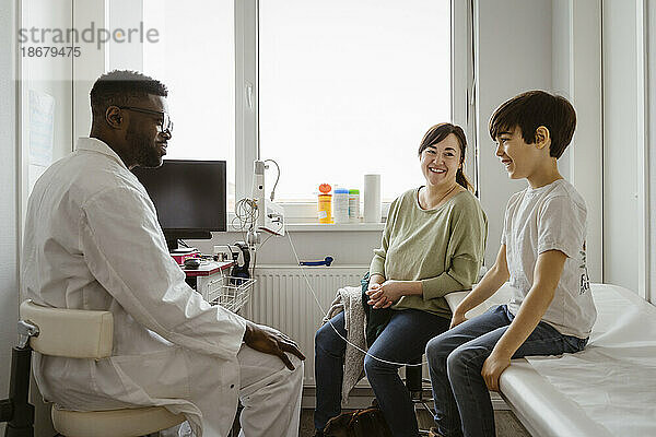 Lächelnder Junge mit seiner Mutter im Gespräch mit einem männlichen Kinderarzt im Untersuchungsraum eines Gesundheitszentrums