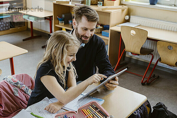 Lächelnde Schülerin  die einen Tablet-PC benutzt  der von einem männlichen Lehrer im Klassenzimmer gehalten wird