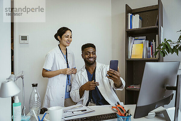 Lächelnde männliche und weibliche medizinische Kollegen führen einen Videoanruf über ein Smartphone in einer Klinik.