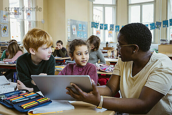 Lehrerin  die einem Schüler und einer Schülerin mit einem digitalen Tablet im Klassenzimmer hilft