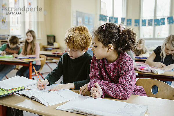 Mädchen sitzt neben einem blonden Jungen  der am Schreibtisch im Klassenzimmer in ein Buch schreibt