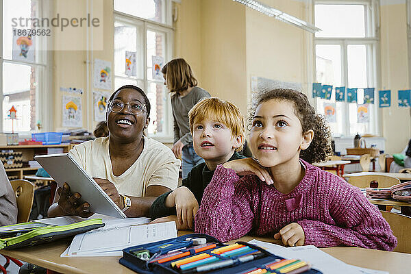 Lächelnder Lehrer mit Tablet-PC  der mit einem Schüler und einer Schülerin am Schreibtisch sitzt  während er im Klassenzimmer nach oben schaut