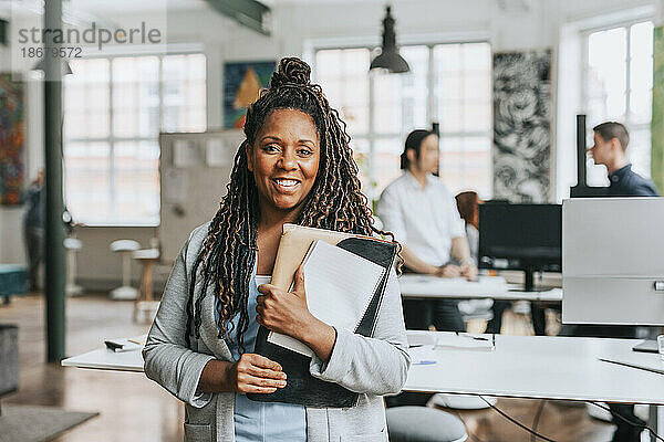 Porträt einer lächelnden Geschäftsfrau mit geflochtenem Haar  die im Büro Akten hält
