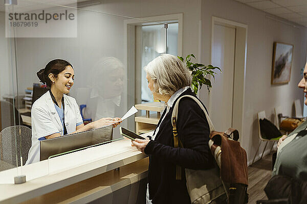 Lächelnde Empfangsdame erklärt dem Patienten das Ausfüllen von Formularen durch einen transparenten Schild in der Klinik